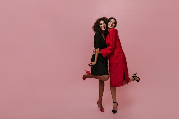 Belle femme moderne en robe midi rouge vif et talons foncés étreignant son amie souriante avec des cheveux bouclés et des vêtements noirs