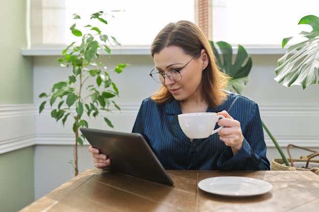 Belle femme mûre en pyjama, lecture de nouvelles, réseaux sociaux, courrier en tablette numérique. Portrait du matin d'une femme d'âge moyen à la maison à table avec une tasse de thé, petit-déjeuner.
