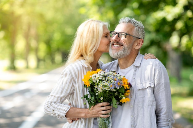 Belle femme mûre tenant des fleurs et embrassant son mari pendant une promenade dans le parc