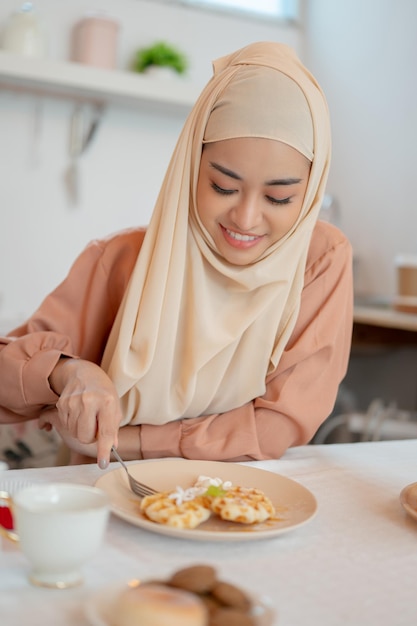 Une belle femme musulmane asiatique mangeant des gaufres prend son petit déjeuner à une table dans la cuisine