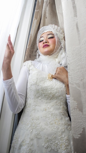 Une belle femme musulmane asiatique pose dans une robe de mariée blanche avec un voile près de la fenêtre à l'air heureux