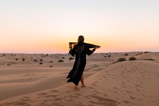 Photo belle femme mystérieuse en robe longue noire arabe traditionnelle se dresse dans le désert au coucher du soleil