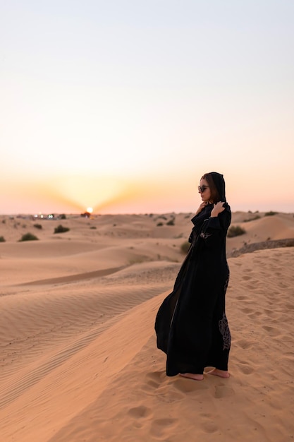 Photo belle femme mystérieuse en robe longue noire arabe traditionnelle se dresse dans le désert au coucher du soleil