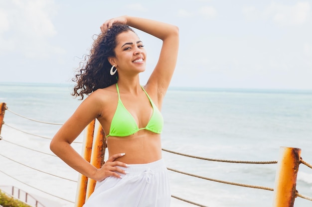 Belle femme noire afro portant un bikini brésilien latino-américain avec son dos souriant dans une station balnéaire regardant les vagues de la mer de l'océan s'écraser