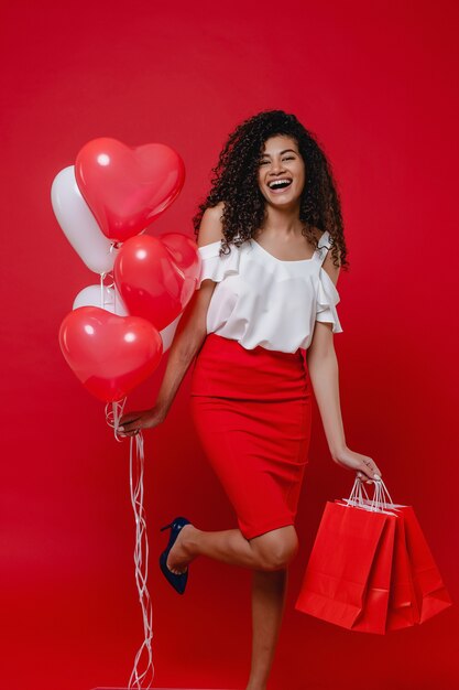 Belle femme noire avec des ballons en forme de coeur coloré et des sacs à provisions sur mur rouge