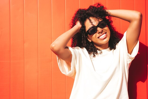 Belle Femme Noire Avec Des Boucles Afro Coiffuremodèle Hipster Souriant En T-shirt Blanc Et Short Sexy Femme Insouciante Posant Dans La Rue Près Du Mur Rouge à Lunettes De Soleil Enthousiaste Et Heureux