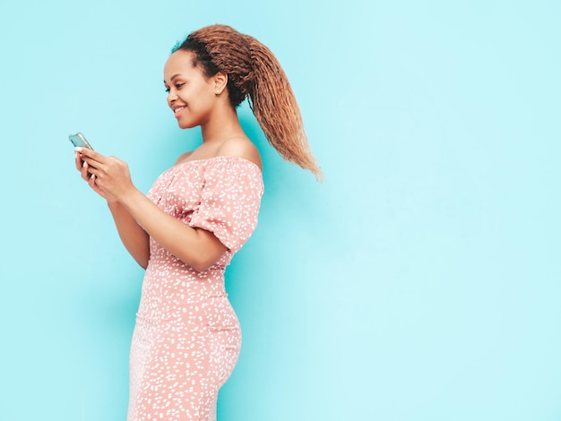 Belle femme noire avec une coiffure afro curlsModèle souriant dans des vêtements d'été à la mode Sexy femme insouciante posant près d'un mur bleu en studioUtilisant des applications pour smartphoneRegardant l'écran du téléphone portable