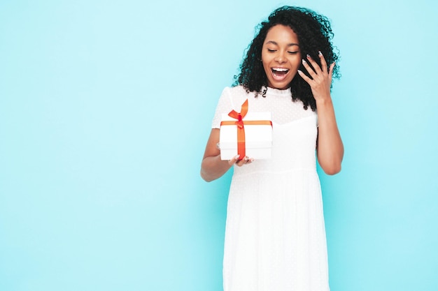 Belle femme noire avec une coiffure de boucles afro Modèle souriant vêtu d'une robe d'été blanche Sexy femme insouciante posant près d'un mur bleu en studio Bronzé et joyeux Tenant une boîte-cadeau Isolé