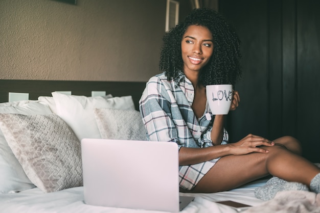 Belle femme noire sur lit avec ordinateur portable et tasse de café