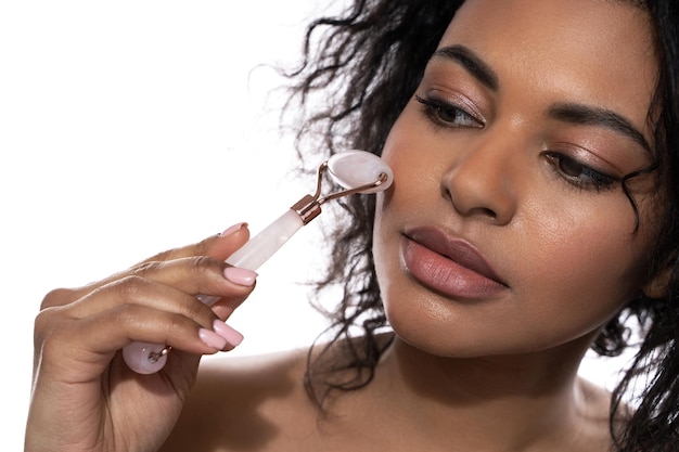 Belle femme noire avec une peau lisse à l'aide d'un rouleau derma quartz rose sur fond blanc