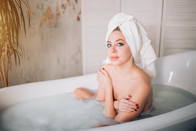 Belle femme nue avec une peau parfaite, des yeux expressifs, des lèvres charnues, une manucure rouge assise dans une baignoire avec une serviette blanche sur la tête dans un salon de spa moderne. Concept de personnes, de bien-être et de soins du corps.