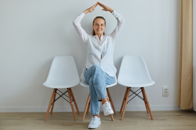 Belle femme optimiste assise sur une chaise à l'intérieur, vêtue d'une chemise blanche et d'un jean, faisant le toit avec les mains au-dessus de sa tête, se sent en sécurité, exprimant son bonheur.