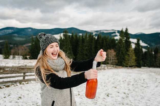 Belle femme ouvre une bouteille de champagne contre les montagnes d'hiver.