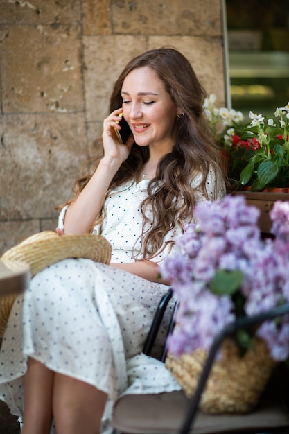 Belle femme parlant sur smartphone près de la fleur de lilas. Modèle mignon et fleurs. Aromathérapie et concept de printemps. Fille au café. Asseyez-vous avec un panier de lilas dans les mains. Fleuriste