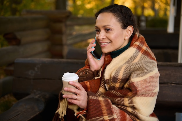 Belle femme parlant sur un téléphone portable, tenant une tasse en carton de boisson chaude, se tenant au chaud tout en étant assise sur un banc en bois enveloppé dans une couverture en laine à carreaux beige confortable par une fraîche journée d'automne