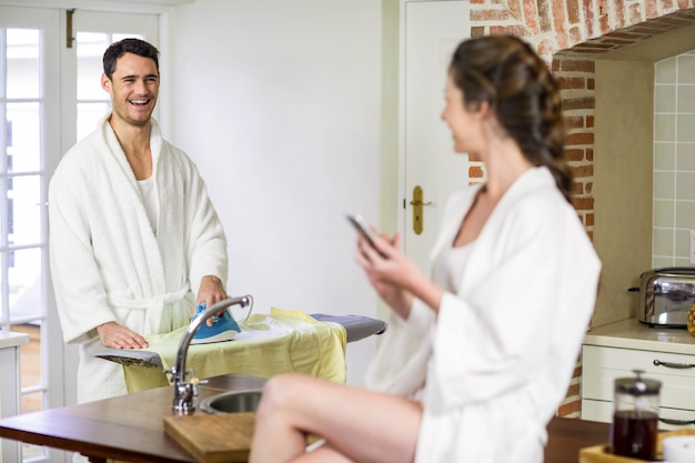 Belle femme en peignoir assis sur le plan de travail de la cuisine et en tapant un message texte sur smartphone pendant qu&#39;un homme repassant des vêtements derrière elle