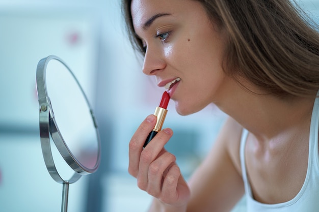 Belle femme peint les lèvres avec du rouge à lèvres rouge à l'aide d'un petit miroir rond pendant le maquillage à la maison le matin