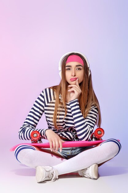 Belle femme pensive assise avec une planche à roulettes et écoutant de la musique sur fond rose