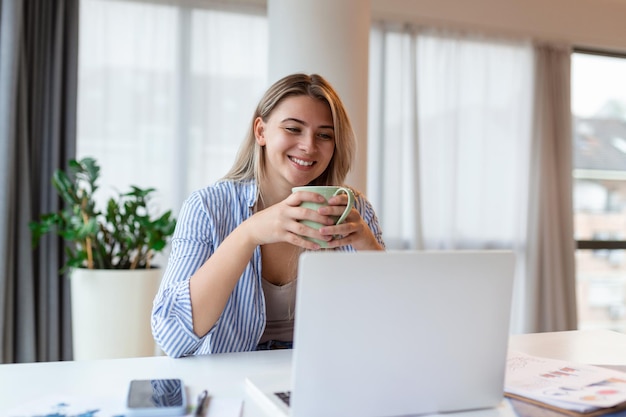 Belle femme pigiste notant des informations pour la planification d'un projet faisant un travail à distance via un ordinateur portable femme riant en lisant des e-mails sur un ordinateur portable moderne