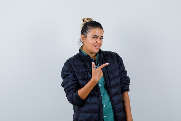 Belle femme pointant vers la droite avec l'index en chemise verte, veste noire et l'air confiant, vue de face.