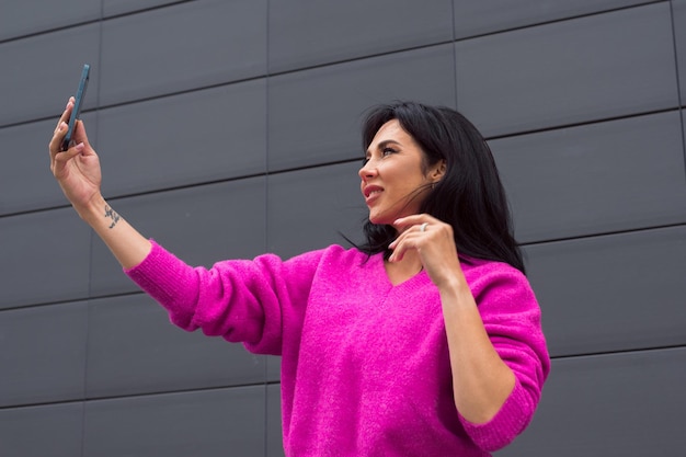 Belle femme portant un pull rose vif posant dans la rue de la ville avec un smartphone