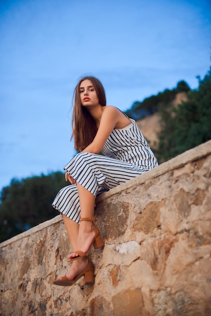 Belle femme portant une salopette rayée et des sandales élégantes assis sur un mur de pierre.