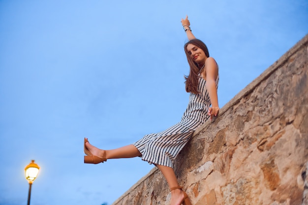 Belle femme portant une salopette rayée et des sandales élégantes assis sur un mur de pierre.