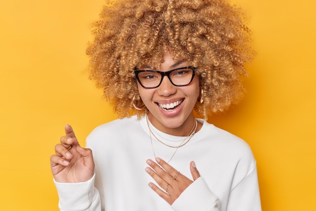 Une belle femme positive aux cheveux blonds bouclés sourit exprime joyeusement des sentiments sincères garde la main sur la poitrine porte des lunettes et le cavalier blanc se sent heureux isolé sur fond jaune.