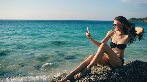 Belle femme prenant selfie à l'aide de téléphone sur la plage souriant et appréciant le style de vie de voyage