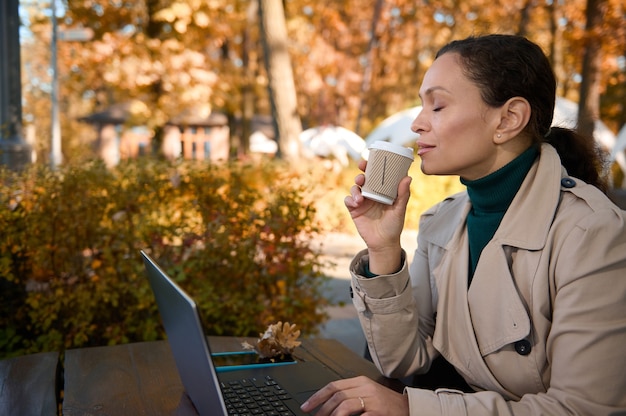 Belle femme profitant d'un travail à distance, se relaxant dans la cafétéria en bois de la chênaie et buvant un délicieux café chaud tout en travaillant sur un ordinateur portable loin de l'agitation de la ville