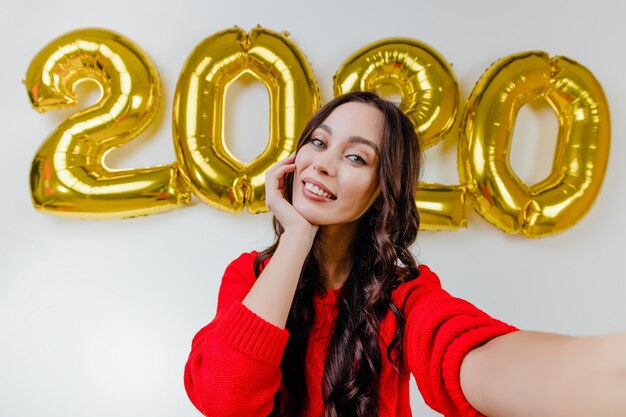 Belle femme en pull rouge faisant selfie drôle devant des ballons de nouvel an 2020