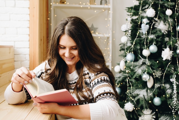 Belle femme de race blanche lisant un livre violet près de l'arbre de Noël décoré