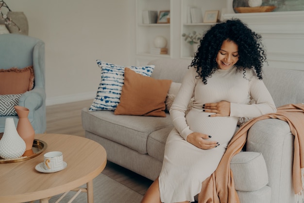 Belle femme de race mixte enceinte souriante assise sur un canapé et touchant son ventre