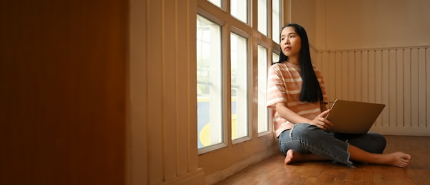 Belle femme regardant à travers les fenêtres tout en utilisant un ordinateur portable qui met ses genoux et assis sur le plancher en bois du salon.