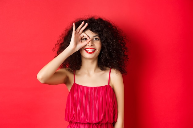 Belle femme regarde à travers le signe correct avec excitation, vérifiant l'offre promotionnelle, debout en rouge sur fond rouge.