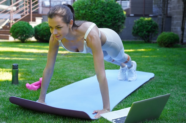 Belle femme de remise en forme faisant un exercice de planche en regardant des didacticiels en ligne sur un ordinateur portable, s'entraînant en plein air. Mode de vie sain. La fille fait du sport au jardin.