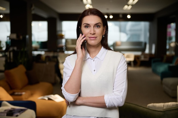 Belle femme responsable des ventes confiante dans un magasin de meubles parle sur un téléphone portable en regardant la caméra