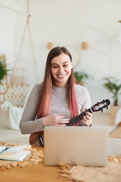 Une belle femme rieuse apprend à jouer du ukulélé à distance. L'enseignant donne une leçon de ukulélé en ligne.