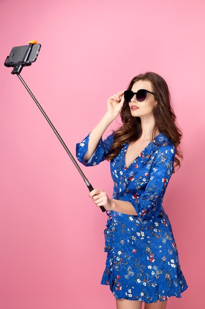 Belle femme en robe bleue avec des fleurs, des lunettes de soleil prenant selfie en studio sur rose