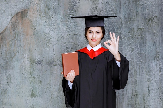 Belle femme en robe de graduation posant avec un livre et donnant le signe ok. Photo de haute qualité