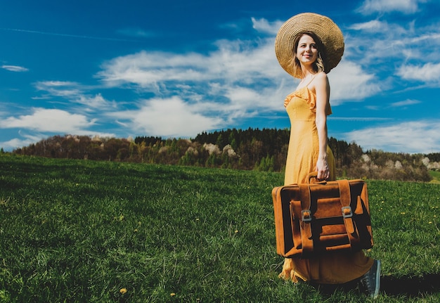 Belle femme en robe jaune et valise sur la prairie de montagne avec des pissenlits