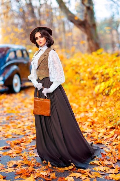 Belle femme en robe vintage, chemisier en dentelle et chapeau avec voile debout près d'une voiture marron rétro