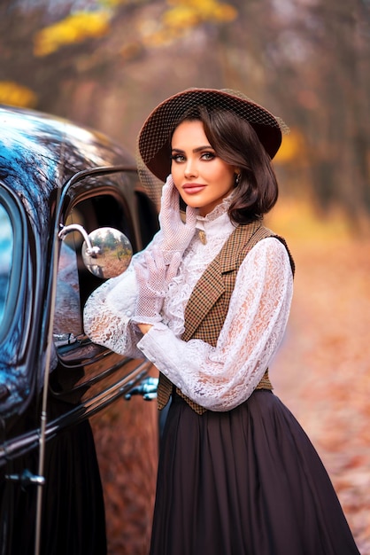 Belle femme en robe vintage, chemisier en dentelle et chapeau avec voile debout près d'une voiture marron rétro