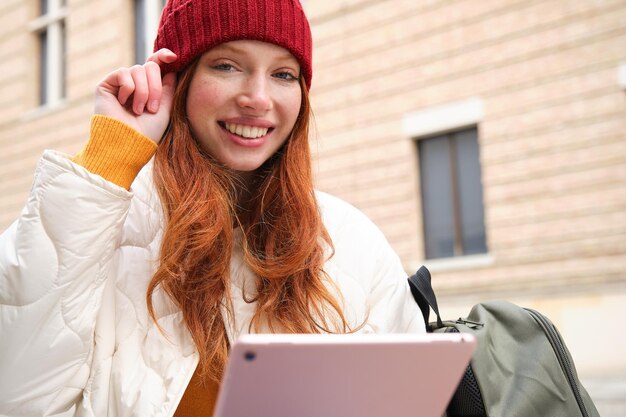 Belle femme rousse au chapeau rouge est assise avec un sac à dos et un thermos à l'aide d'une tablette numérique à l'extérieur conn