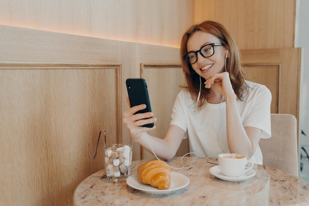 Belle femme rousse dans les écouteurs assis dans un café et faisant un appel vidéo sur téléphone mobile