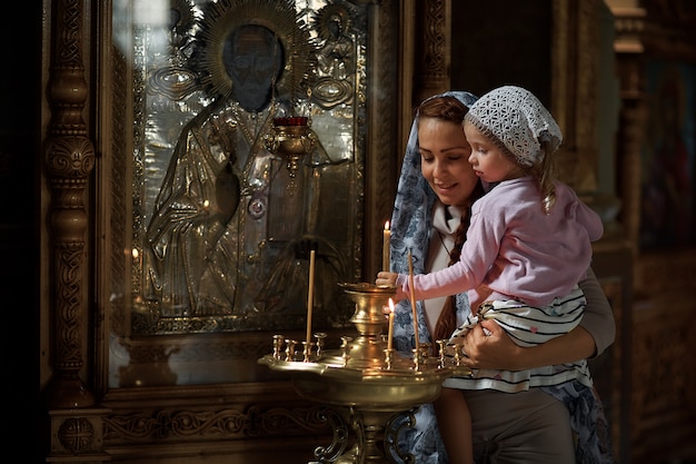 Belle femme russe dans une écharpe et aux cheveux rouges tenant une petite fille et allume une bougie devant une icône dans l'église orthodoxe russe.