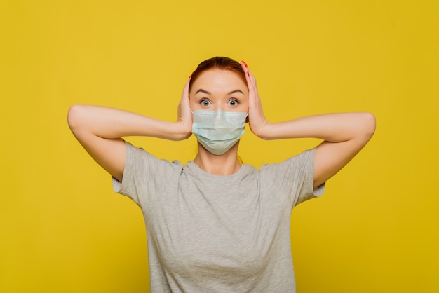 Une belle femme s'accroche à sa tête et est choquée par la propagation du coronavirus. Belle fille rousse dans un masque de protection sur un mur jaune. Le concept de protection et de prévention