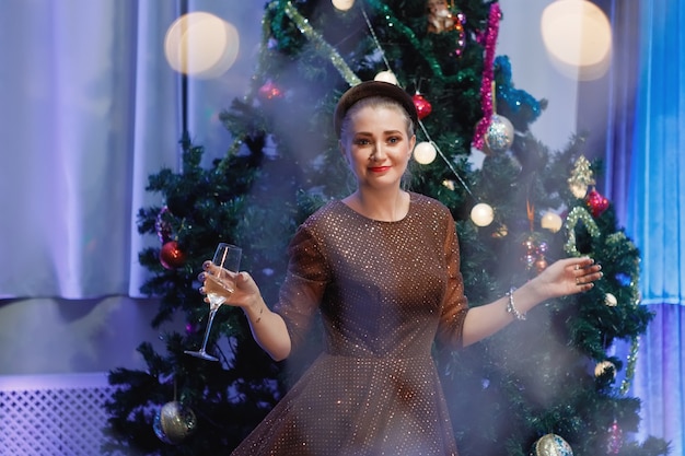 Belle femme s'amusant avec une coupe de champagne près d'un arbre de Noël. Une femme rit, sourit, pose. Filtre spécial bruit et grain vintage, lumières floues.