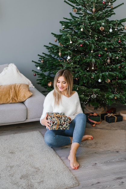 La belle femme s'assied sur le plancher près de l'arbre de Noël dans le salon, tenant une surprise