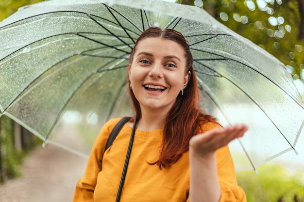 Une Belle Femme Se Tient Sous Un Parapluie Transparent Et Tend La Main Pour Attraper Des Gouttes D'eau...